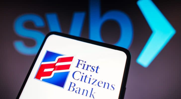 First Citizens Bank (FCNCA) logo displayed on smartphone in front of SVB logo in dark background behind phone