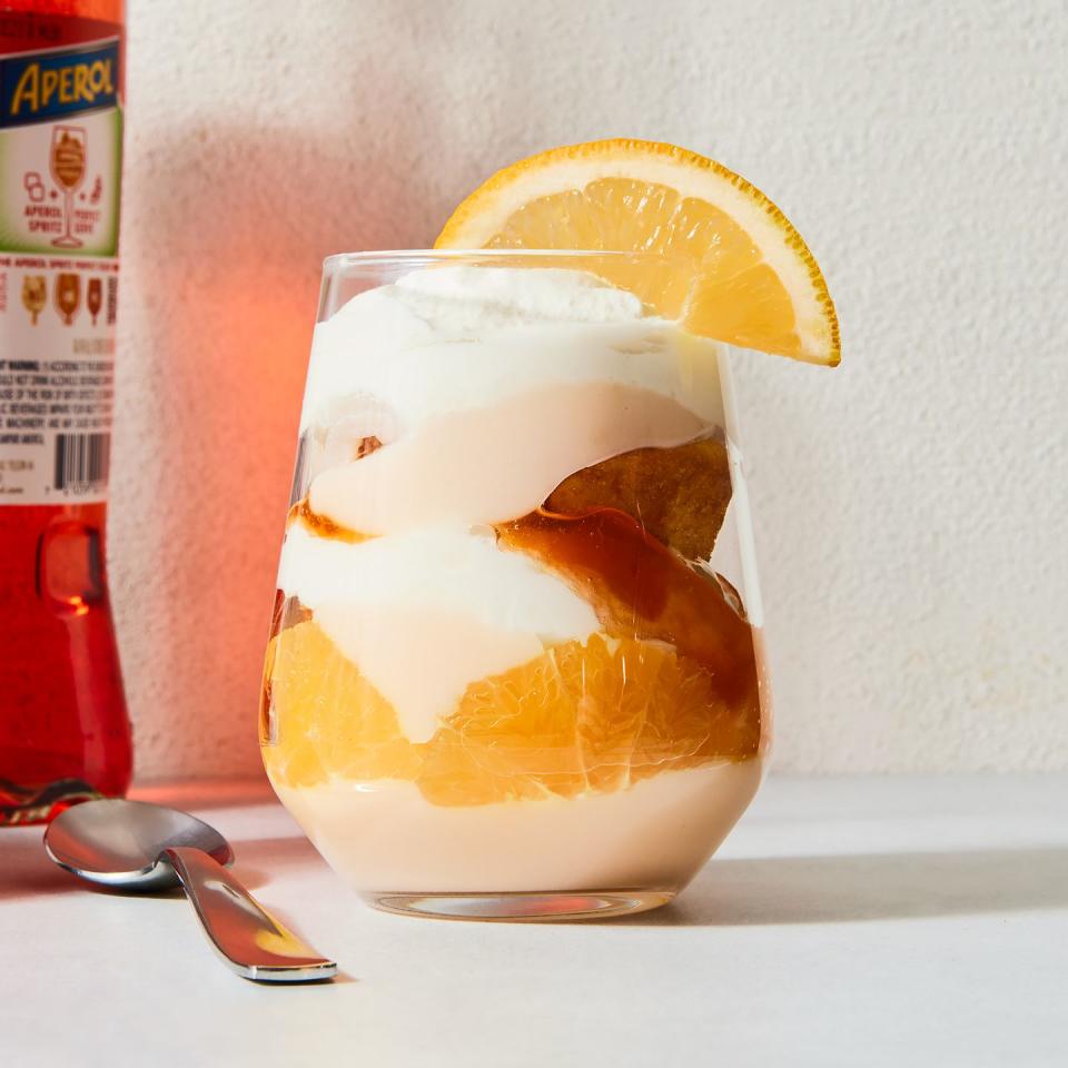 a stemless wine glass filled with vanilla cake, whipped cream, aperol spiked jam, and pudding with an orange slice on the rim