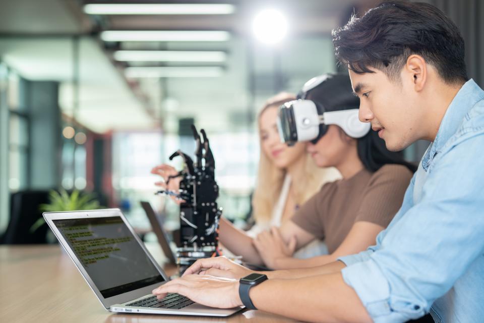 A group of people sitting at a table in an open office, working on laptops and using VR headsets.