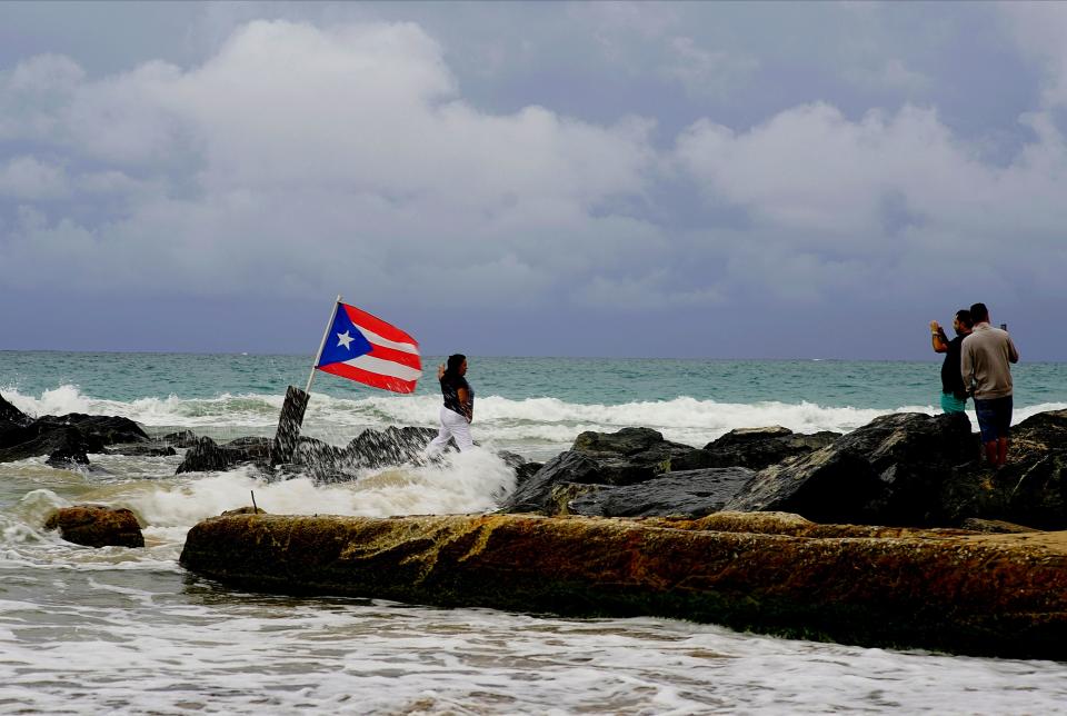 A woman poses for a photo backdropped by ocean waters and a Puerto Rican national flag, after the passing of Tropical Storm Dorian, in the Condado district of San Juan, Puerto Rico, Wednesday, Aug. 28, 2019.