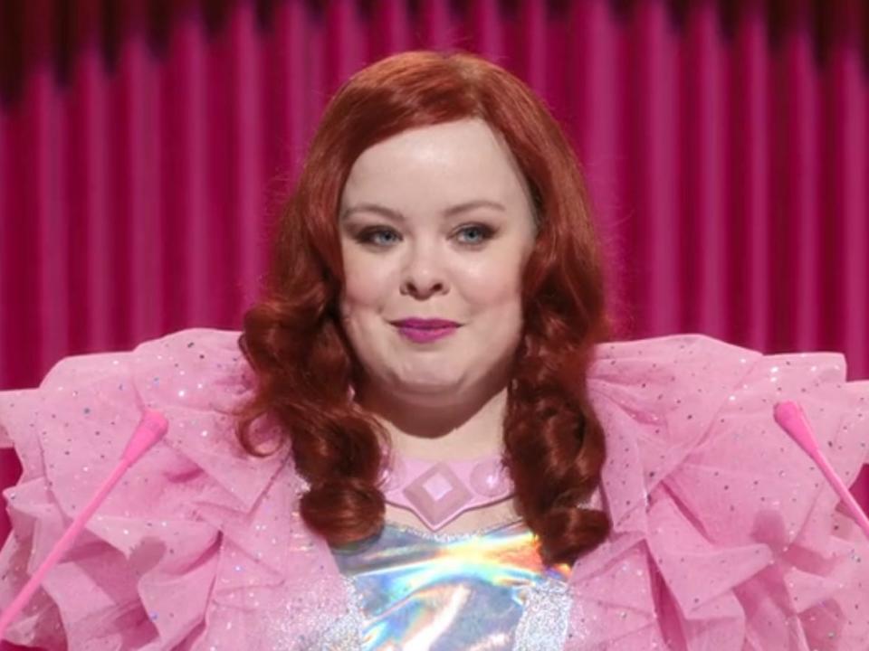 nicola coughlin in barbie with red hair and a frilly pink and silver dress, standing at a lectern