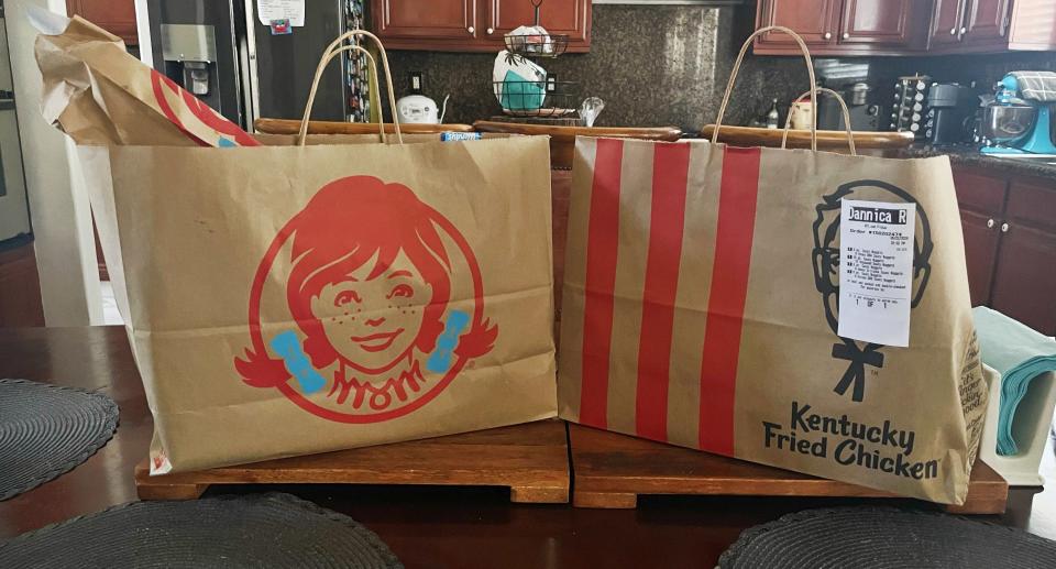 Two large takeout bags, one from Wendy's and one from Kentucky Fried Chicken, are placed on a kitchen island with wooden cabinets and a refrigerator in the background