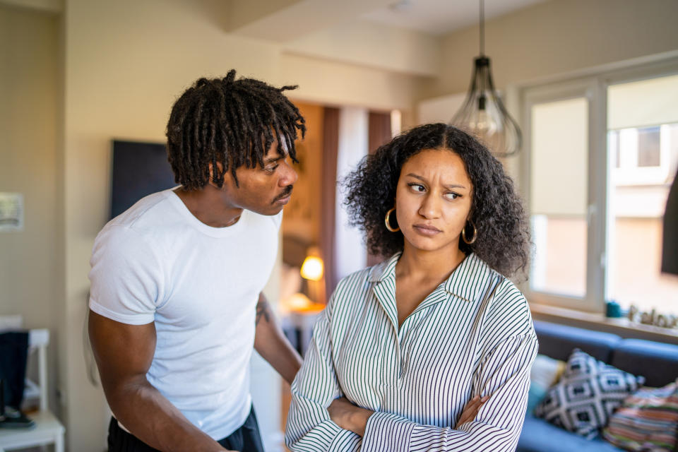 A man and a woman are standing in a living room. The man appears to be speaking intently, while the woman looks away with a furrowed brow and crossed arms