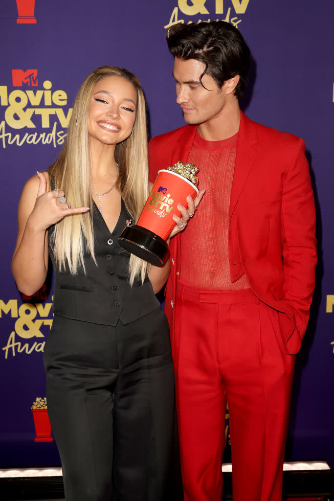 Madelyn Cline in a vest and pants and Chase Stokes in a suit with sheer shirt, posing with an award at the MTV Movie & TV Awards. Madelyn holds the award and smiles