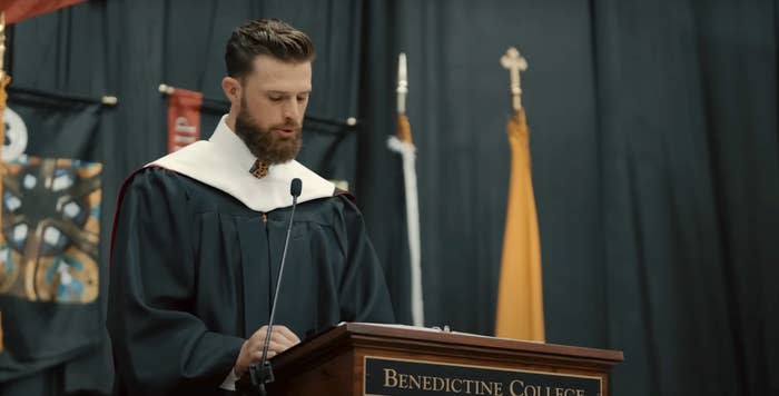 A man in graduation robes speaks at a podium labeled "Benedictine College" at a ceremony