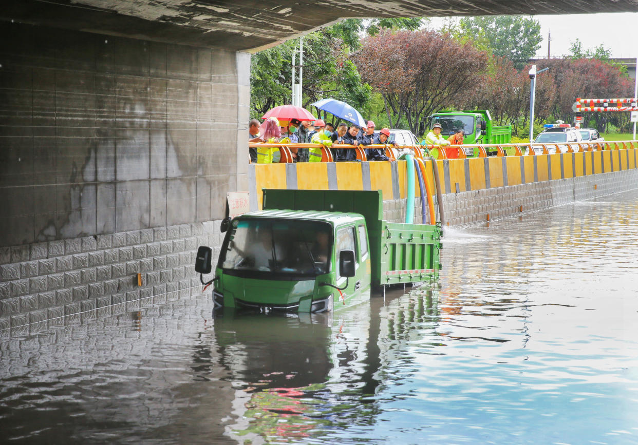 A small green dumptruck is stranded in high water below a bridge while pedestrians with umbrellas stand and watch.