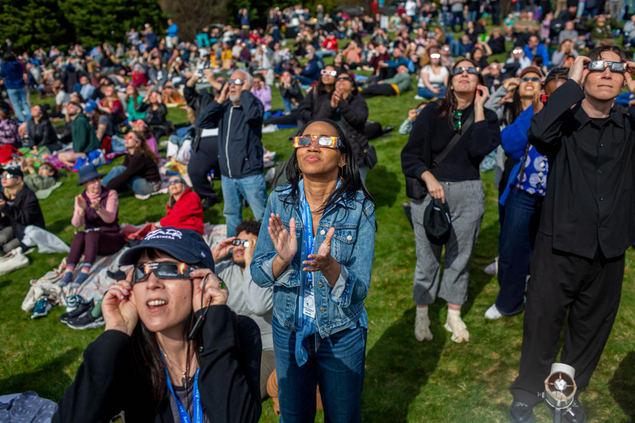 Several hundred people on a grassy hill wear eclipse glasses while looking up.