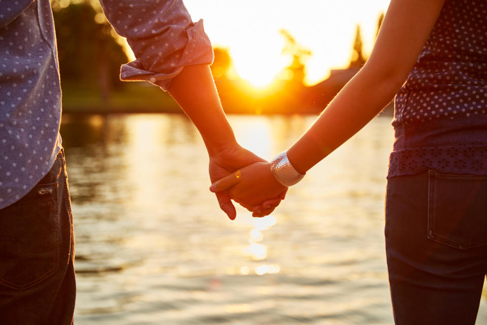 Two people holding hands near a waterfront at sunset, facing away from the camera. Their clothing features casual polka dot patterns