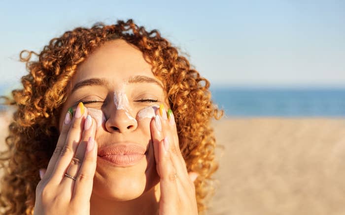 A person with curly hair is applying sunscreen to their face while standing on a sandy beach with the ocean in the background