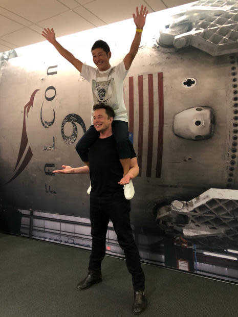 Musk, dressed in black, smiles while holding Japanese space tourist Yusaku Maezawa on his shoulders. In the background is a previously flown Falcon 9 rocket. / Credit: SpaceX