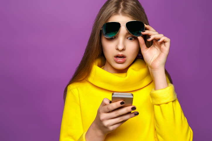 A woman in a yellow sweater shocked as she looks at her phone.