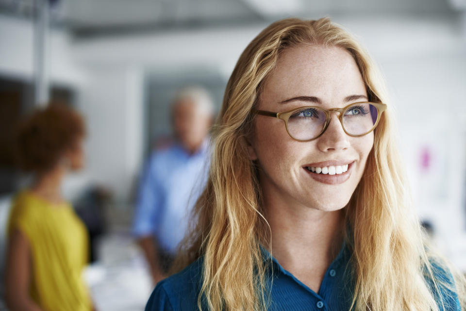 Thoughtful businesswoman smiling while looking away at creative office with colleagues in background