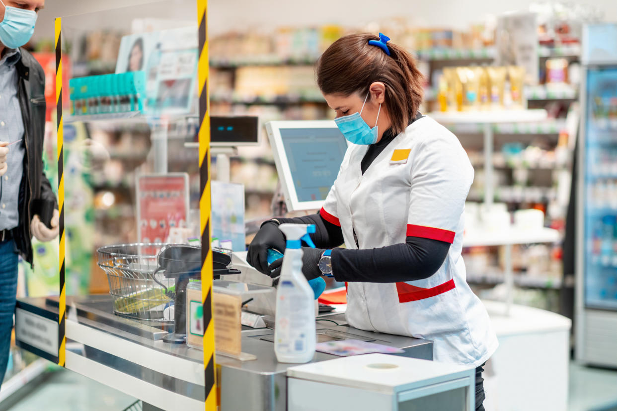 Female sales clerk behind acrylic wall scanning disinfection items for price check, customer waiting to pay, both following safety protocol amid Covid-19 outbreak