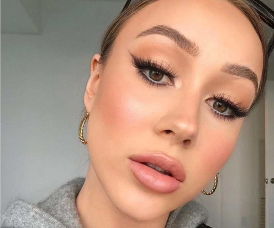 A caucasian woman wears a full face of makeup with winged black eyeliner infront of a pale grey background.