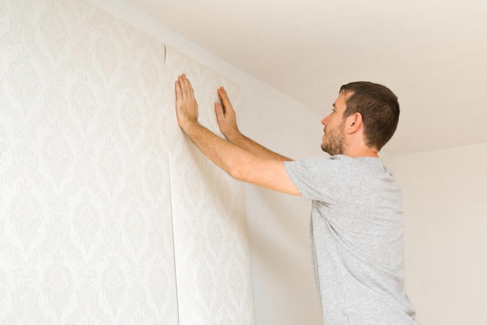Young adult man holding new wallpaper high on a white wall. 