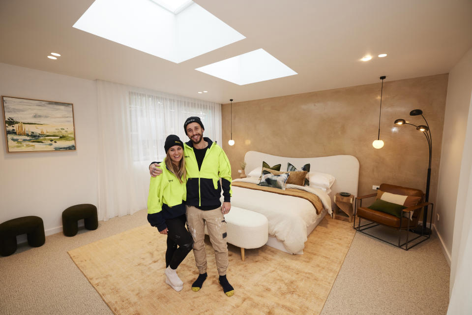 Kristy and Brett smile posing in the centre of their room, in front of a large bed and a cream coloured rug. 