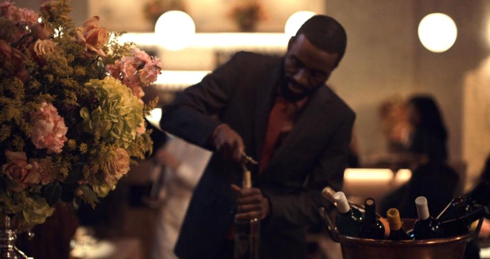 A man in formal attire opens a bottle of wine at an upscale event, with flowers and other guests in the background