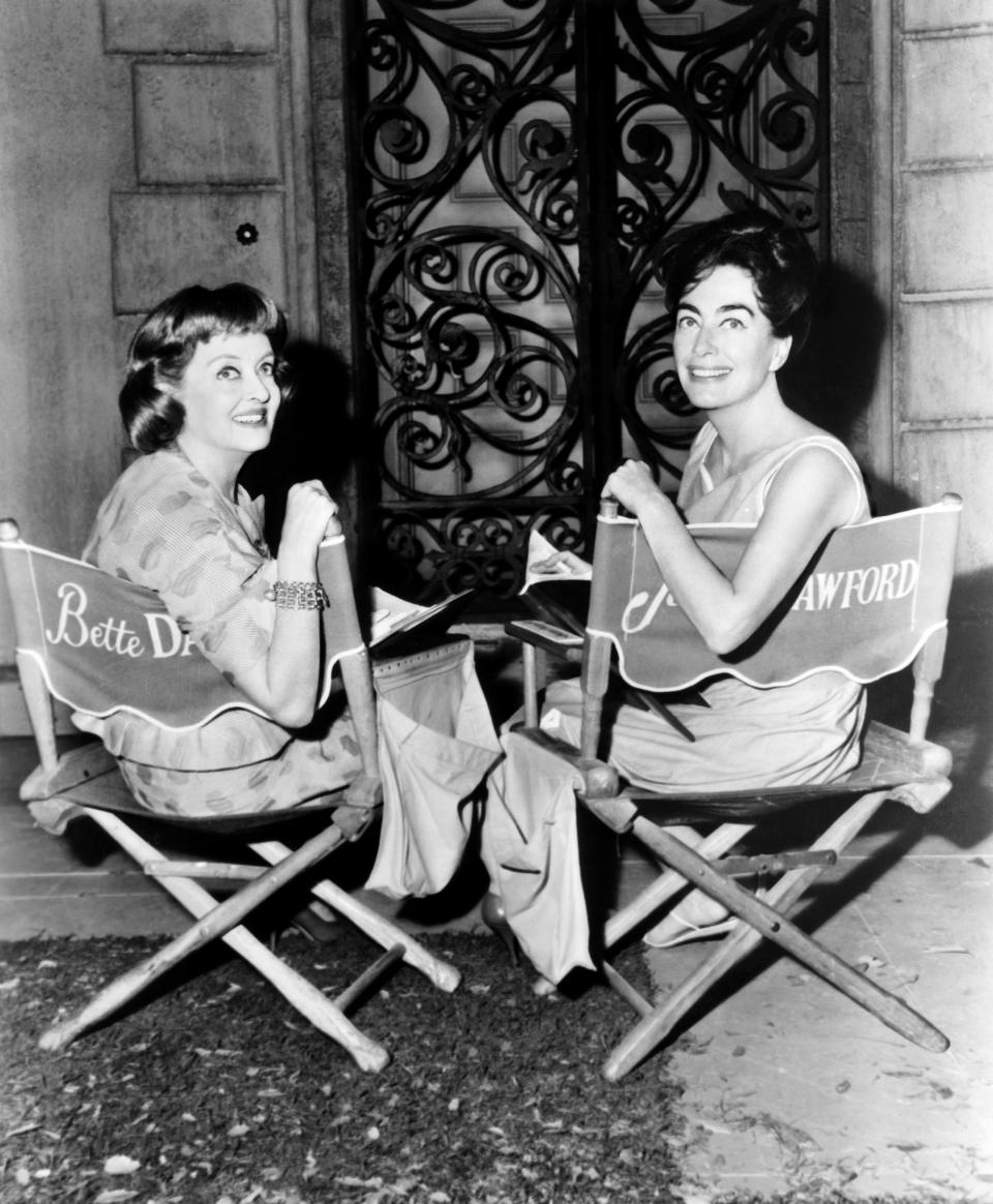 Bette Davis and Joan Crawford are seated in director’s chairs with their names on the backrests, turned slightly towards the camera, smiling on a film set