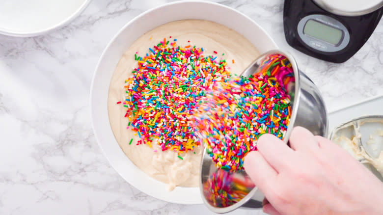 Person pouring rainbow sprinkles into cake batter.