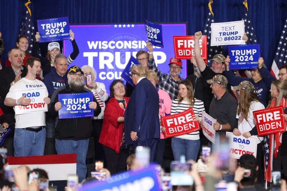 People hold signs that say Trump 2024 and stand together, as a man wearing a dark suit faces drowsed them.