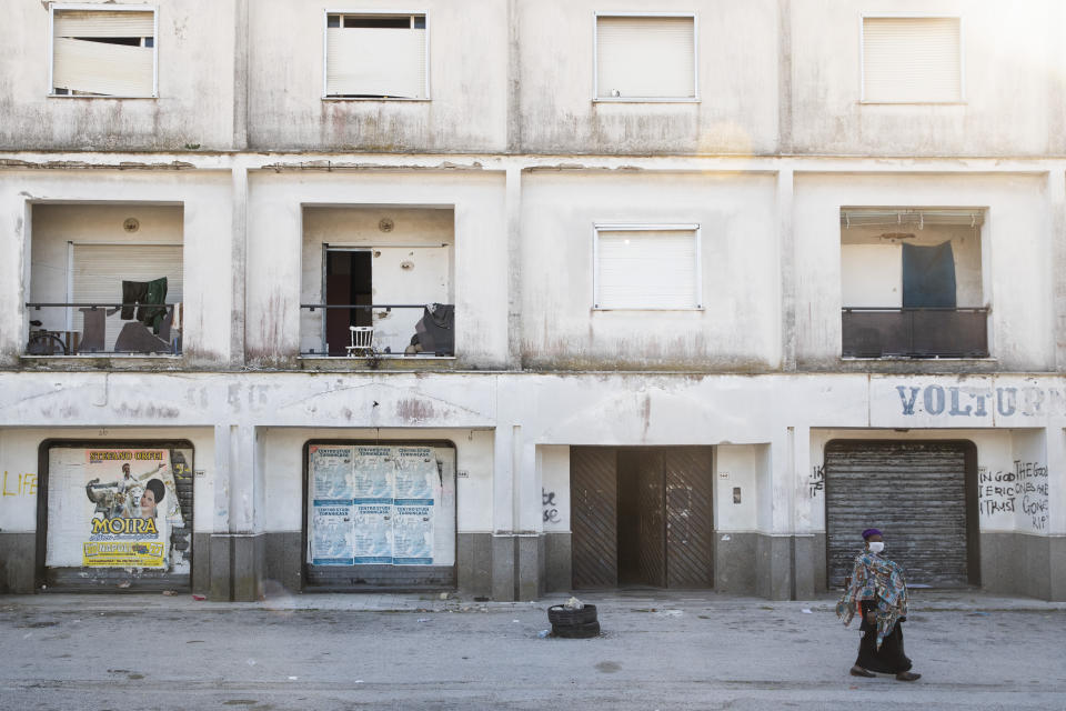 In this photo taken on Monday, April 27, 2020, a woman wearing a sanitary mask to protect against COVID-19 walks past a building in Castel Volturno, near Naples, Southern Italy. They are known as “the invisibles,” the undocumented African migrants who, even before the coronavirus outbreak plunged Italy into crisis, barely scraped by as day laborers, prostitutes and seasonal farm hands. Locked down for two months in their overcrowded apartments, their hand-to-mouth existence has grown even more precarious with no work, no food and no hope. (AP Photo/Alessandra Tarantino)