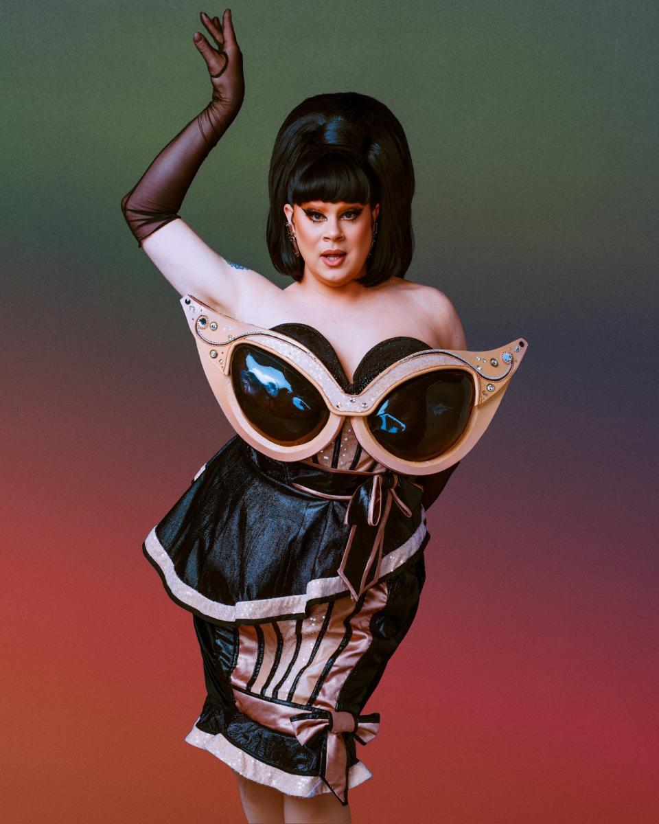 Nina West in an avant-garde, futuristic black and beige dress with large, exaggerated glasses as part of the outfit poses with one arm raised