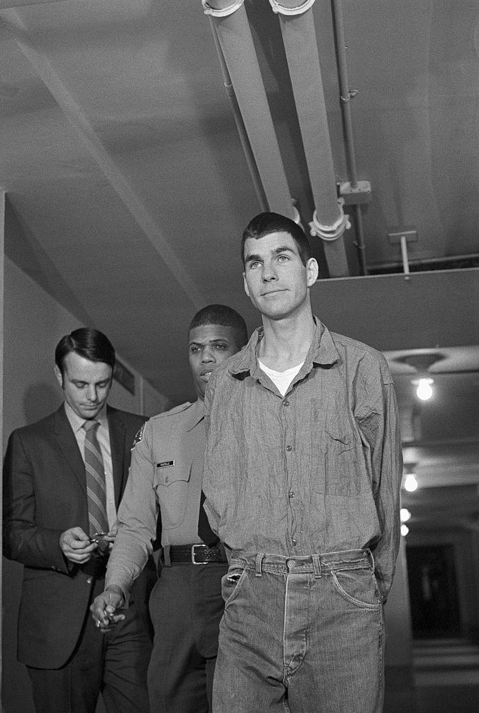 a man wearing a buttoned shit is led while handcuffed down a hallway, escorted by two people