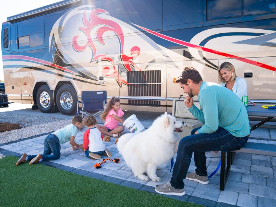 A family with a dog sitting next to a parked Class A RV.