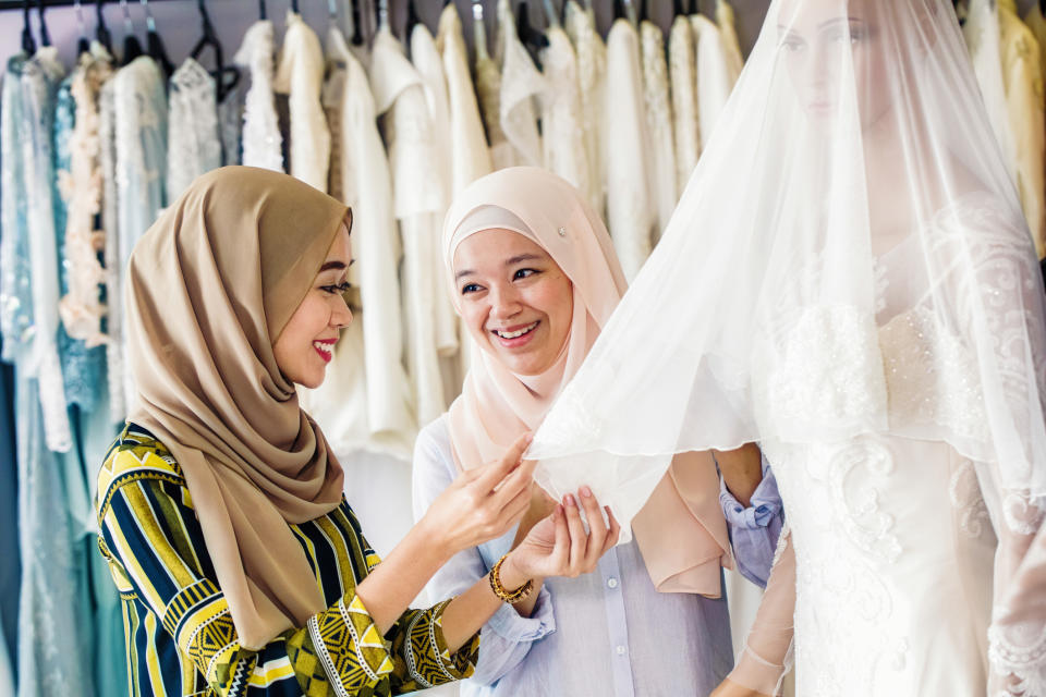 Two women, one in a striped hijab and another in a pale hijab, smile while adjusting a wedding gown on a mannequin. Several wedding dresses hang in the background