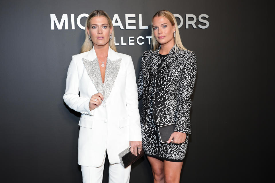 Identical twins Amelia Spencer and Eliza Spencer. Amelia at left wears a white over sized pants suit with silver lapels and Eliza wears a black and white printed mini dress and coat. They both carry black clutch purses and wear their long blonde hair loose.