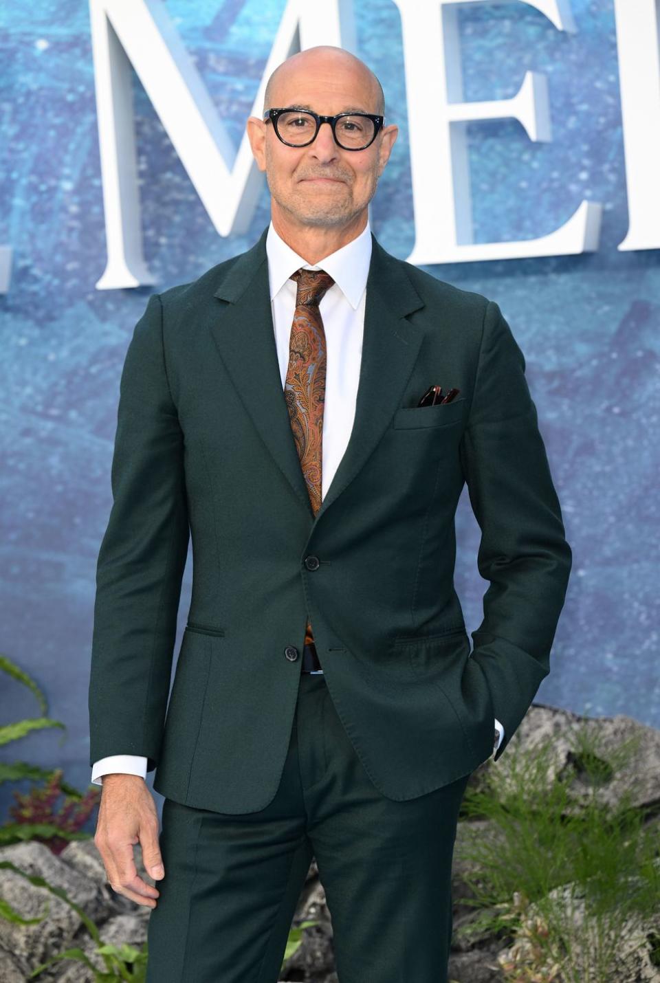 stanley tucci wearing a suit with a brown and orange paisley print tie and glasses on the red carpet at the little mermaid uk premiere