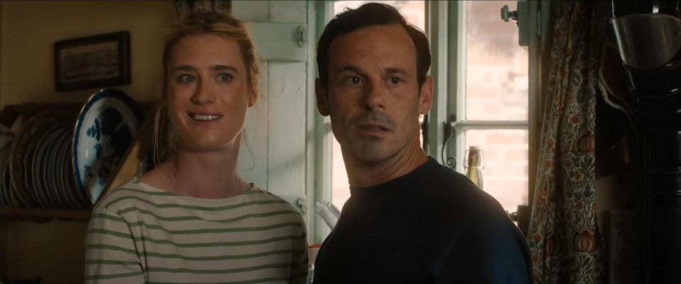 Mackenzie Davis and Scoot McNairy stand together inside a home, looking slightly off-camera with pleasant expressions. Plates are visible in the background