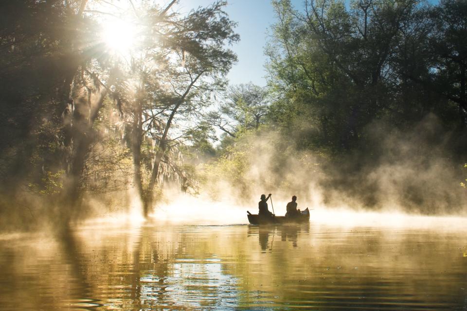 Two people on a canoe paddle through the everglades with sunshine illuminating the mid-afternoon fog around them.
