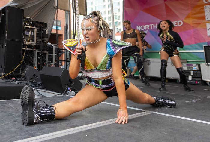 Woman in sparkling futuristic costume performs energetic dance and splits on stage, holding a microphone. Dancers in matching outfits are in the background