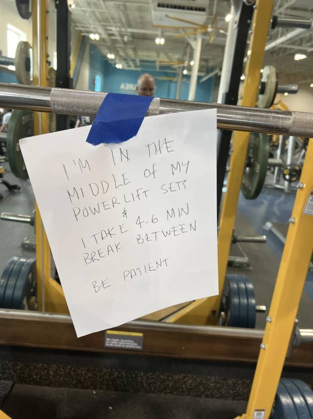 A handwritten sign on a gym barbell reads: "I'm in the middle of my powerlift sets. I take 4-6 min break between. Be patient." A gym-goer is visible in the background