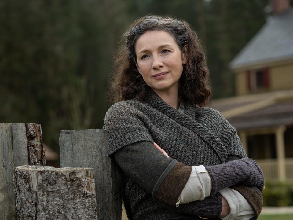 Claire Fraser leaning up against a fence while wearing a knit jumper.