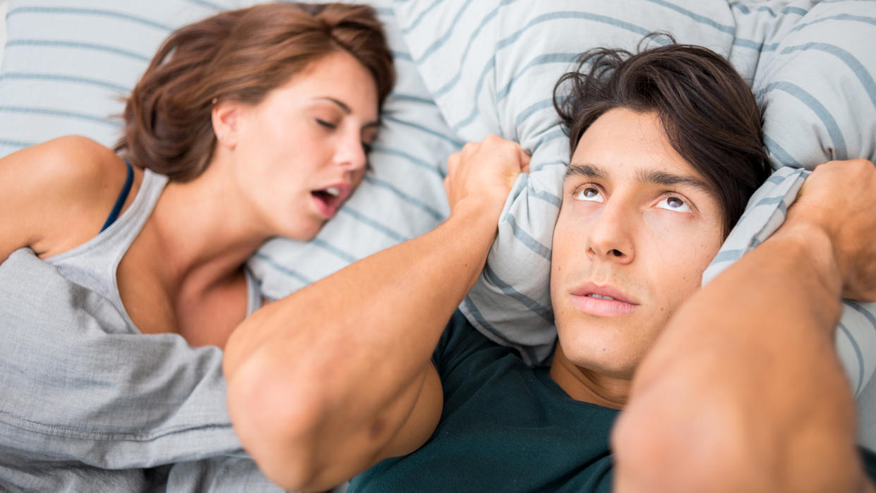  A man with dark hair rolls his eyes in frustration and holds a pillow to his ears because his partner is snoring. 