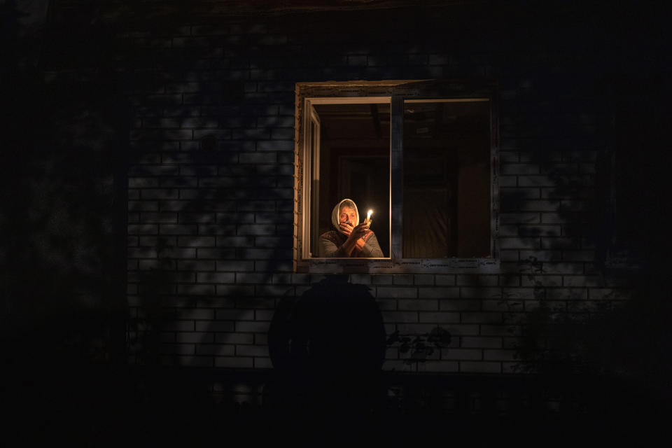 A woman holding a lighted candle, with a shawl over her head, appears illuminated in a window.