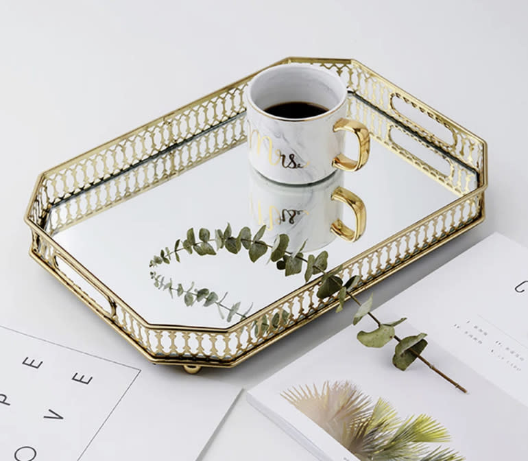 A gold tray holding a cup labeled "Mrs." with coffee, a stem of eucalyptus, and open books nearby