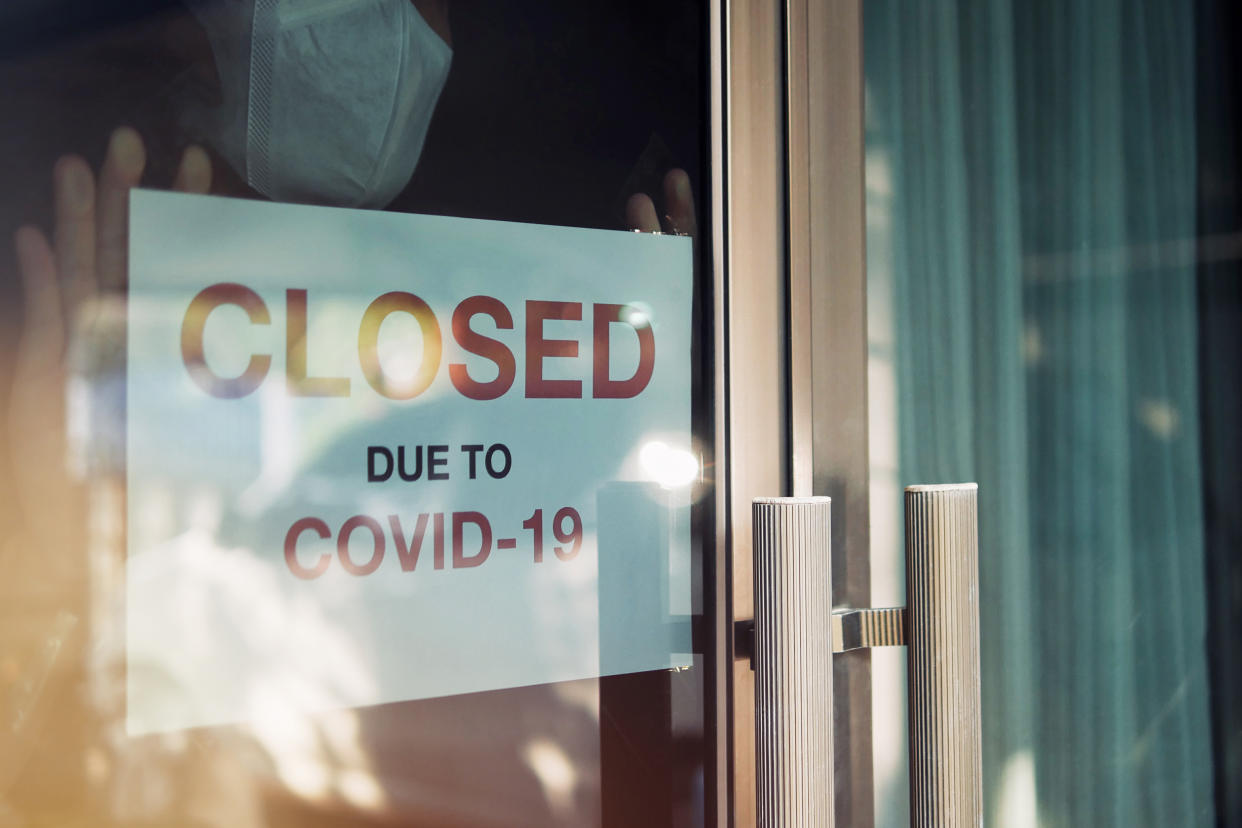 Business office or store shop is closed/bankrupt business due to the effect of novel Coronavirus (COVID-19) pandemic. Unidentified person wearing mask hanging closed sign in background on front door.