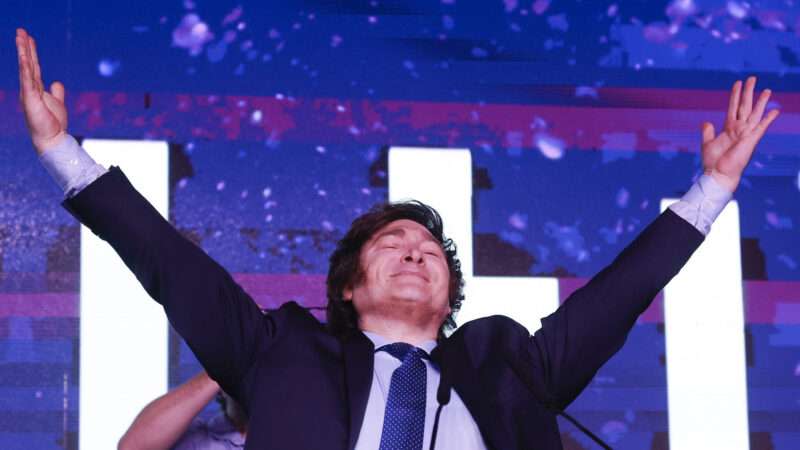 Javier Milei standing with his arms raised in the air in front of a purple background.