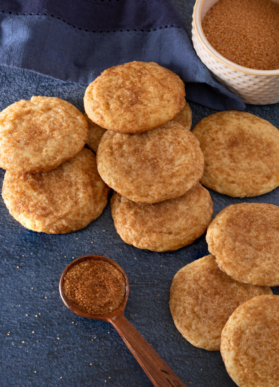 Snickerdoodle cookies are arranged on a textured surface with a wooden spoon and a small bowl of cinnamon sugar nearby