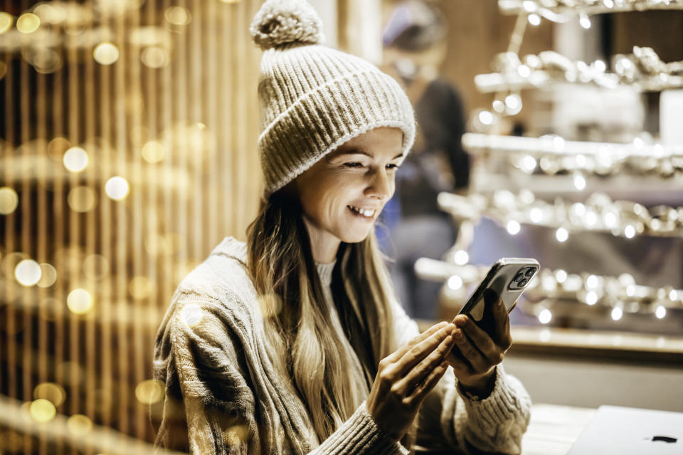 A young American woman using smart phone during festive Christmas shopping, anticipating wonderful family holidays. December ornaments, cafes decorated with fairy lights