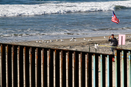 A man holds a U.S. flag while standing on the beach in San Diego, U.S., and behind the border fence between Mexico and the United States, as seen from Tijuana, Mexico November 14, 2018. REUTERS/Jorge Duenes