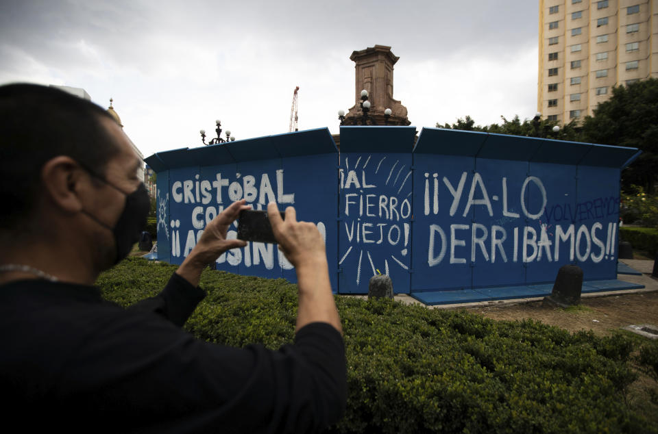 A pedestrian takes a photo of graffiti that reads in Spanish "Christopher Columbus assassin, we've already knocked him down," on a temporary metal barrier set up to protect the perimeter of the Christopher Columbus statue which was removed by authorities on Paseo de la Reforma in Mexico City, Monday, Oct. 12, 2020. Mexico has has a hard time dealing with the legacy of Cristopher Columbus, and officials are still being coy about why the statue was removed over the weekend. (AP Photo/Fernando Llano)