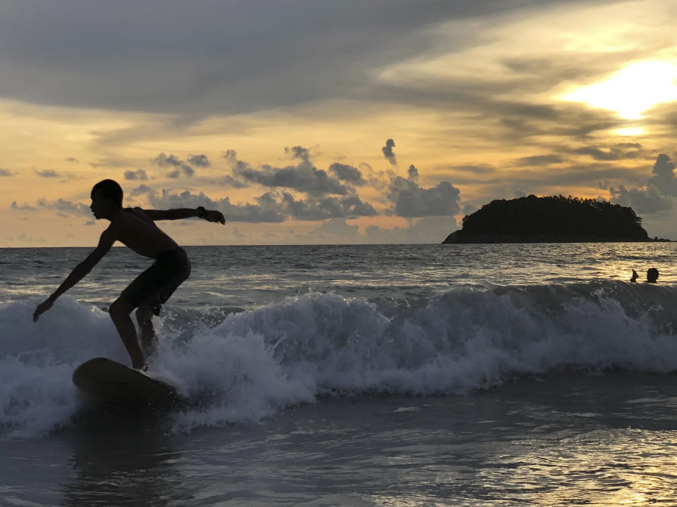 A surfer catches a wave as the sun sets over Kata Beach on the resort island of Phuket, Thailand on Sunday, May 26, 2019. Thailand plans to allow vaccinated foreigners to visit the southern resort island of Phuket without quarantining on arrival in a step toward reviving the country's big but battered tourism industry. (AP Photo/Adam Schreck)