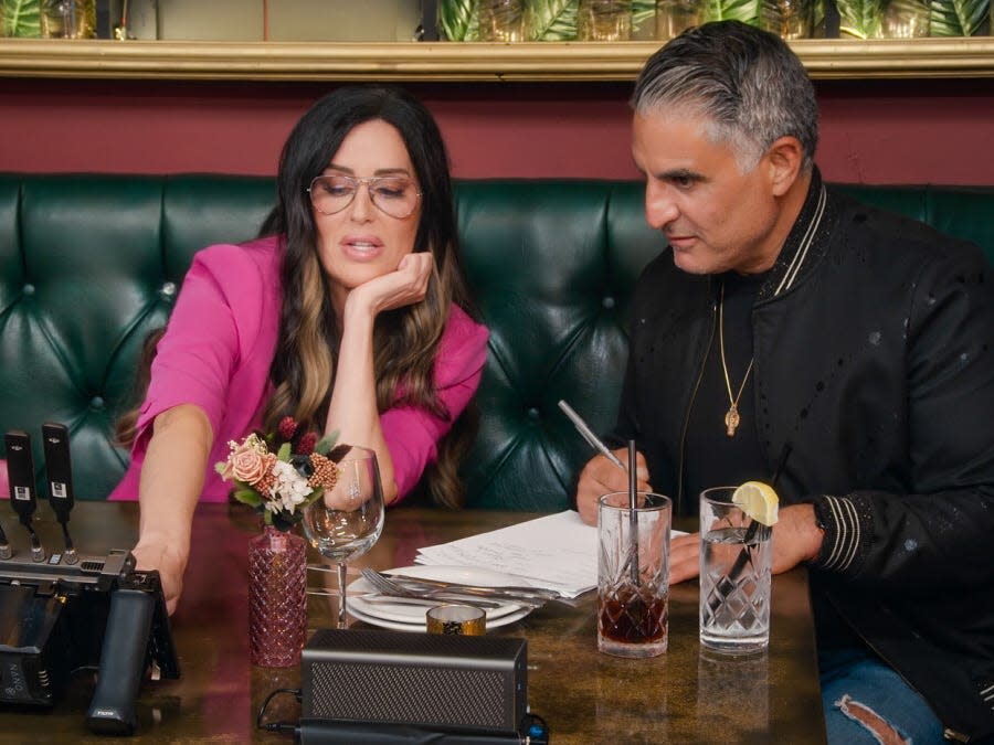 Patti Stanger and Reza Farahan sitting at a table with drinks and a notepad.