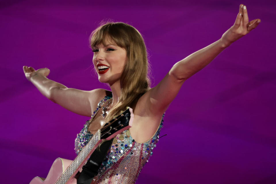 Taylor Swift performing with a guitar, wearing a sequined outfit, arms outstretched