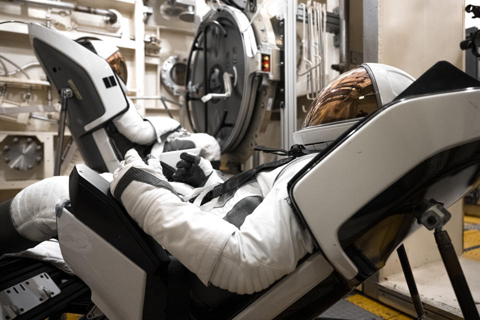 Two astronauts, one in the foreground looking left and the other in the background looking right, are wearing white space suits and leaning back in their white and black bucket suits.