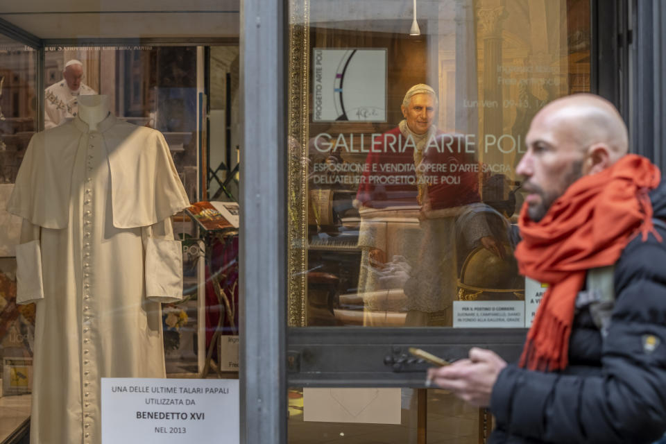 A pedestrian walks by a painting of Pope Emeritus Benedict XVI and one of the last cassocks worn by him before his resignation in 2013, according to the director of the Progetto Arte Poli gallery where it is displayed, near the Vatican, Tuesday, Jan. 3, 2023. The Vatican announced that Pope Benedict died on Dec. 31, 2022, aged 95, and that his funeral will be held on Thursday, Jan. 5, 2023. (AP Photo/Ben Curtis)